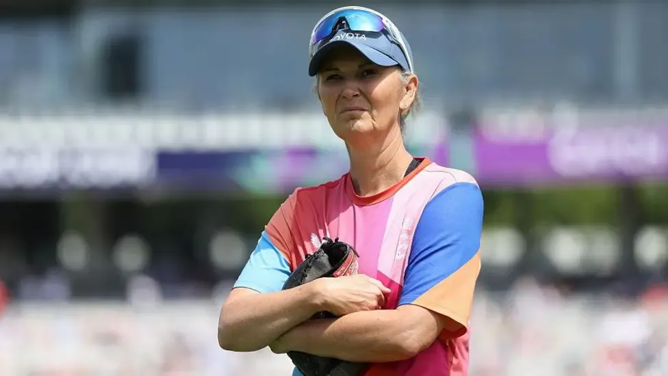 Charlotte Edwards, wearing a blue cap with sunglasses on top and a multicoloured t-shirt, stands with her arms folded