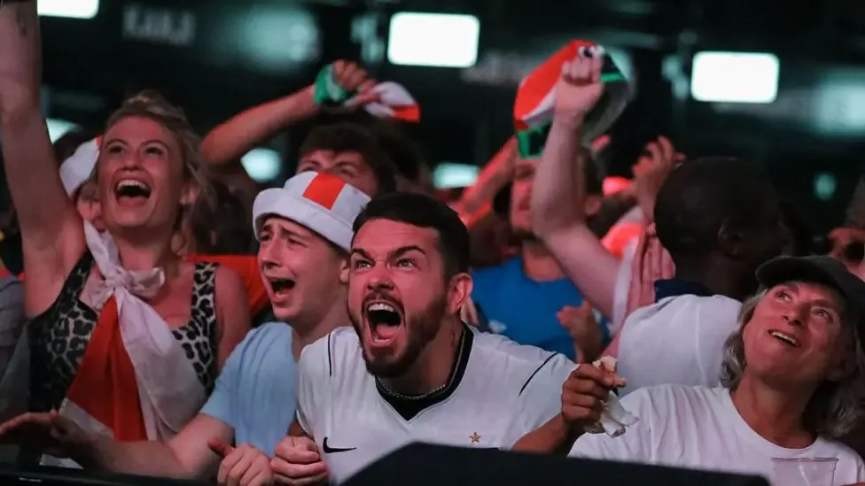 A fan in a white shirt and St George's Cross headband screams with clenched fists at Boxpark Croydon. Other fans around him shout and wave flags.